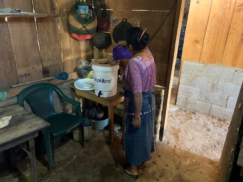 A woman in a pink top and blue striped skirt stands by a wooden table, using a purple pitcher to scoop water into a white Ecofilro bucket in a rustic kitchen.