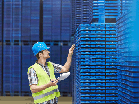 A worker wearing a blue hard hat and high-visibility vest gestures toward stacked blue pallets in a warehouse while holding a clipboard or notebook.