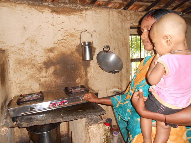 A woman holding a small child watches as she uses a two-burner gas stove in a rustic kitchen with hanging pots on a mud wall.