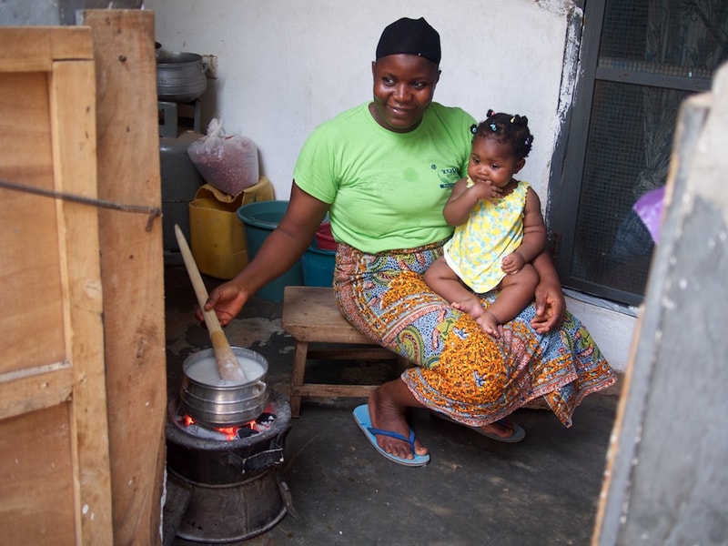 A woman in a bright green shirt and patterned skirt sits on a wooden bench with a small child on her lap, stirring a pot over an open fire in an outdoor kitchen.