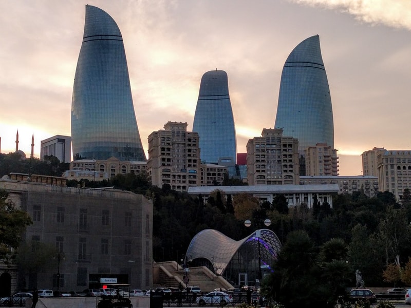 Three tall curved glass skyscrapers rise behind a row of beige buildings, with a modern arched pavilion in a hillside park below during sunset.