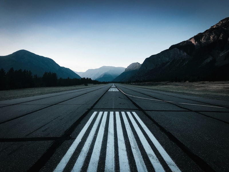 Long runway extending into distant mountains with white parallel markings, flanked by forest and rugged peaks under a clear blue sky.