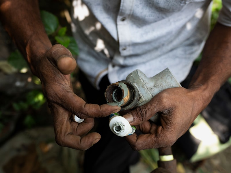 A close-up of dark-skinned hands assembling a metal valve or pipe fitting, with small white threaded parts and a light blue-gray mechanical component held between fingers.