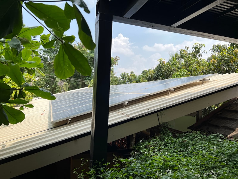 Solar panels installed on a slanted roof next to green foliage and trees, with a dark wooden beam frame in the foreground and a bright blue sky with clouds.