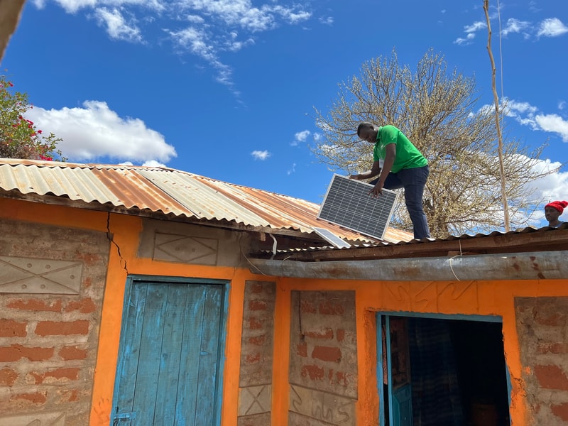 Person on a corrugated roof installing a solar panel, wearing a green shirt; bright blue sky, orange and blue-painted building walls, leafless tree in background.