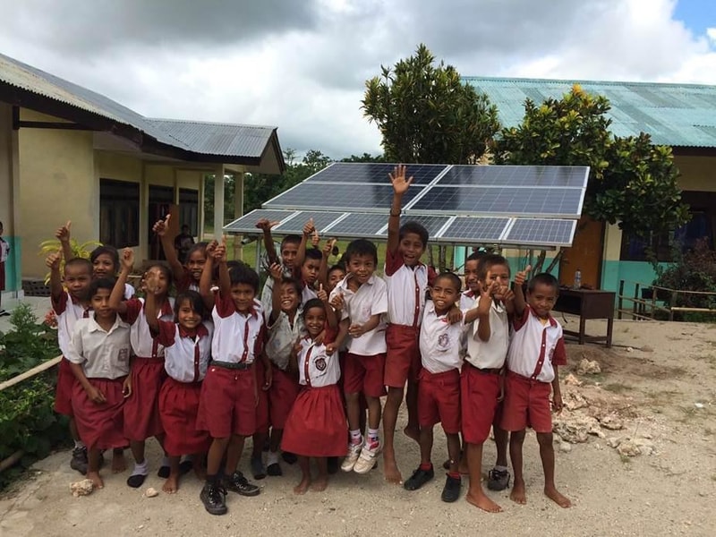 A group of smiling schoolchildren in white shirts and red shorts pose together outdoors, some raising hands, with a large solar panel array and school buildings in the background.