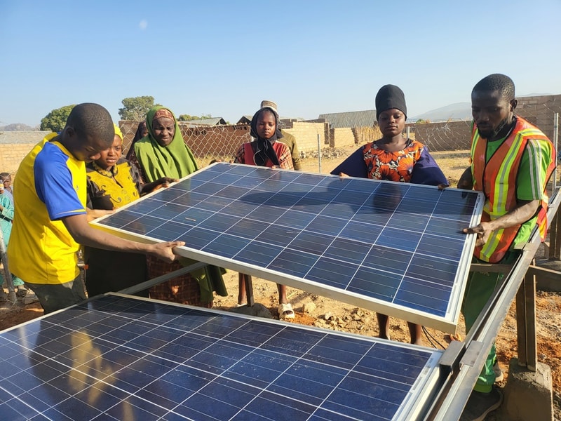 Group of people installing and inspecting large solar panels outdoors in a desert-like area, with a mix of traditional clothing and a safety vest.