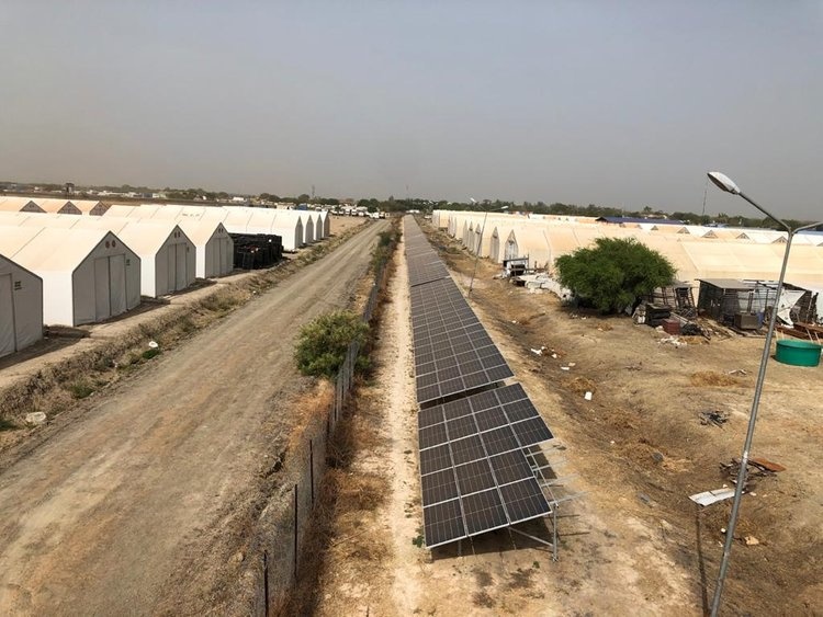 Solar panels line a dirt road between rows of white tents in a temporary camp, with a lamp post, sparse vegetation, and clear sky overhead.