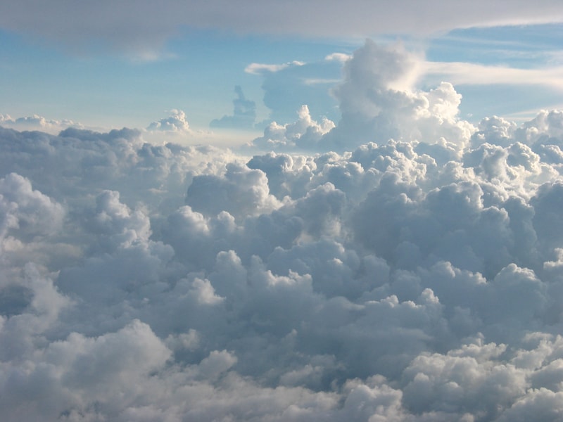 Vast expanse of fluffy white clouds viewed from above, with soft blue sky peeking through and a few towering cumulus formations in the distance.