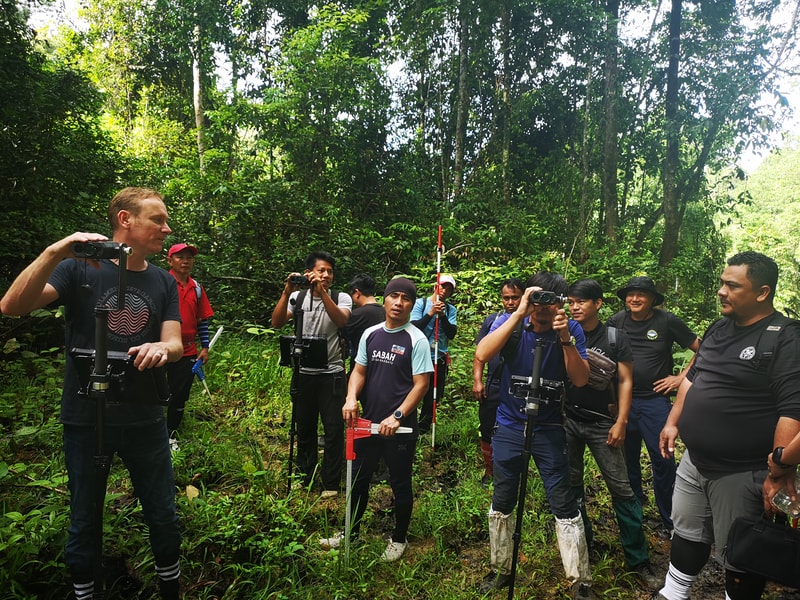 Group of hikers in a dense forest, some with cameras and gear, standing with trekking poles and equipment, focused on an activity or guide in outdoor terrain.