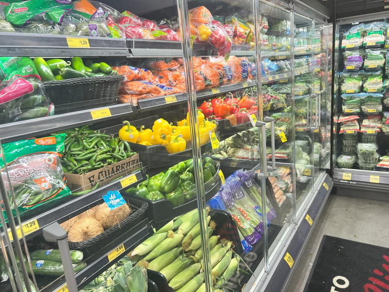 A grocery produce aisle with glass doors displays colorful vegetables including peppers, peppers, cucumbers, carrots, corn, onions, and packaged greens on refrigerated shelves.