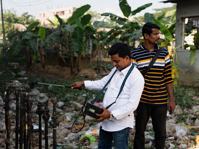 Two men test a water meter with a handheld device outdoors in a yard scattered with trash and banana plants, one operating the gauge while the other watches.