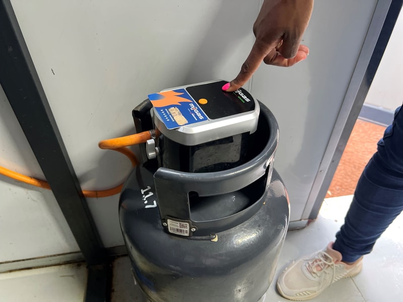 Person pressing a detector on a gray circular device mounted on a ceiling or wall; orange cable runs along surface, sneakered foot and hand visible.