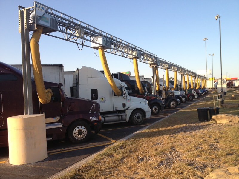 Tractor-trailer trucks lined up in a parking row with yellow flexible exhaust hoses connected to a rooftop fueling/airing gantry under a clear blue sky.