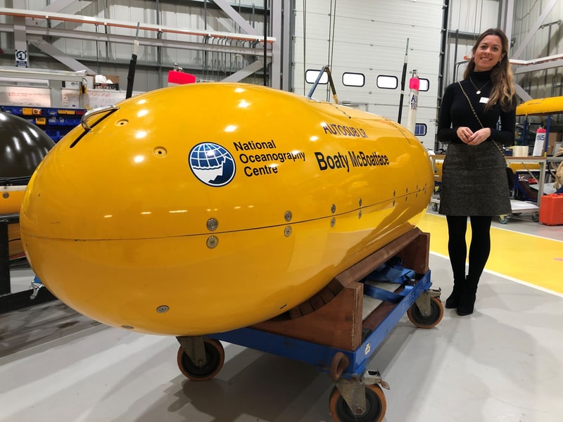 Yellow submarine-shaped buoy with National Oceanography Centre logo, labeled Boaty McBoatface, on a blue wheeled cart inside a workshop; a smiling person stands nearby.