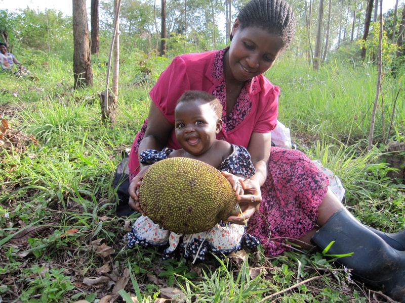 A smiling woman in a pink dress sits in a grassy yard with a small child, both holding a large, spiky jackfruit together.