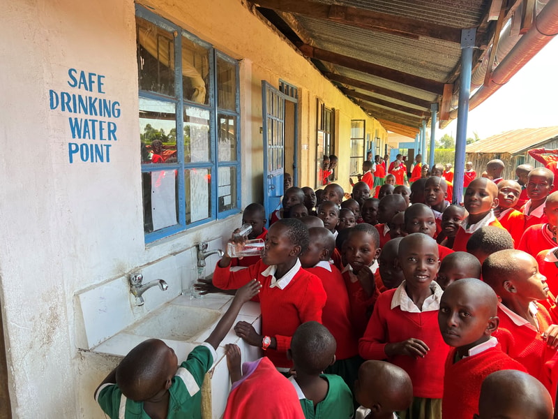 Young students in red uniforms line up at a public water point labeled “SAFE DRINKING WATER POINT,” collecting and drinking water from communal taps outside a building with blue-framed windows.