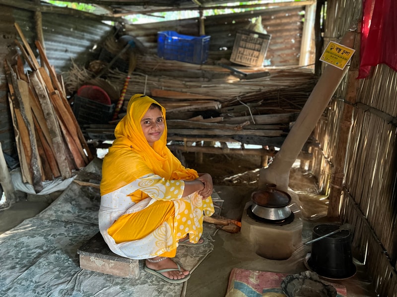 A woman in a bright yellow headscarf and dress sits on a low mat inside a rustic, cluttered shed with wooden planks, a small stove, and cooking pot nearby.