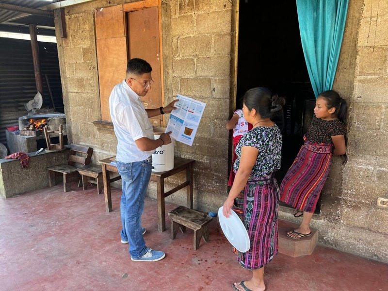 A man in a white shirt and jeans shows a printed flyer to two women outside a rustic brick house; one woman holds a white plate, while a girl stands in the doorway watching.