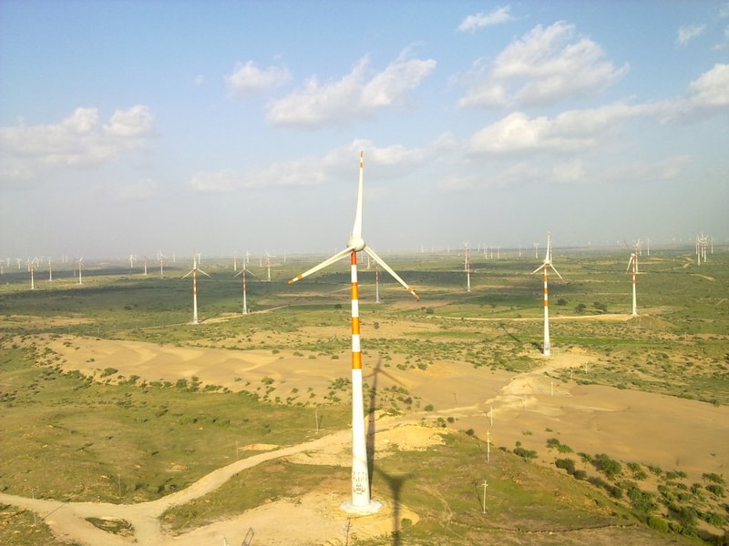 Wide aerial view of a desert landscape with numerous tall wind turbines spread across the flat terrain under a blue sky with scattered clouds.