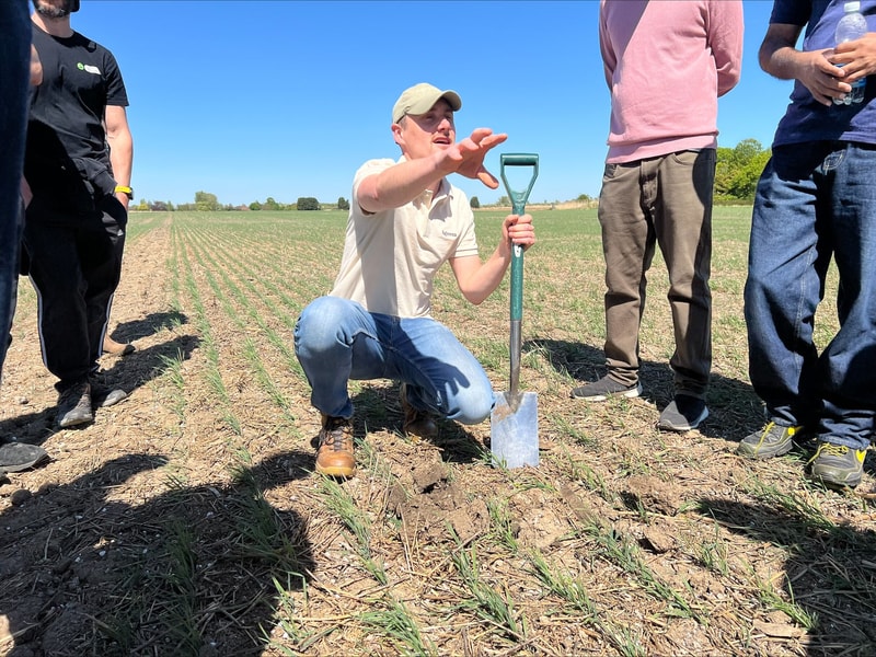 A man kneels in a field with a shovel, gesturing with one hand while others stand around him on a sunny day.