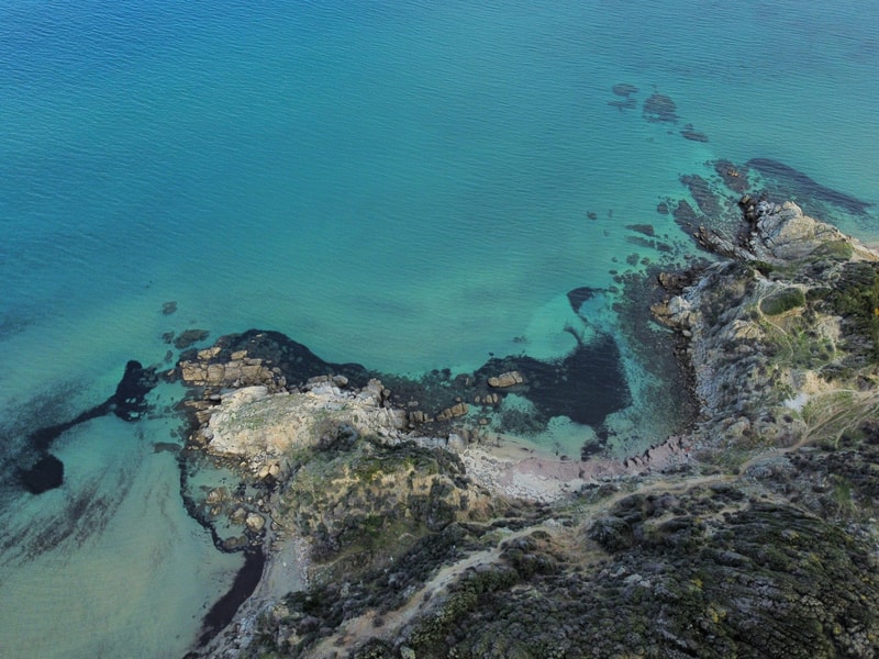 Aerial view of a rocky coastline with turquoise and deep blue ocean, clear shallow waters near the shore, and rugged cliffs with scattered vegetation.