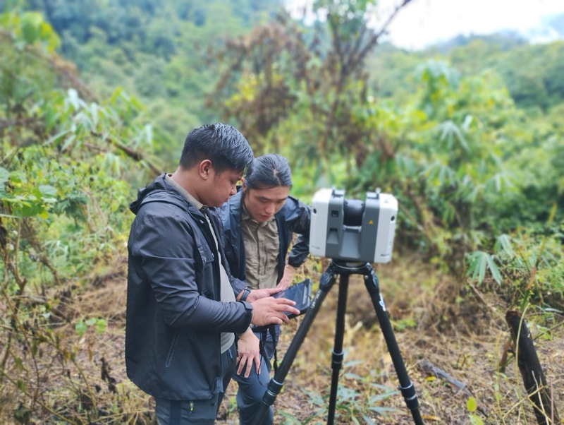 Two people stand in a lush outdoor setting with a large robotic device on a tripod, examining or operating it together.