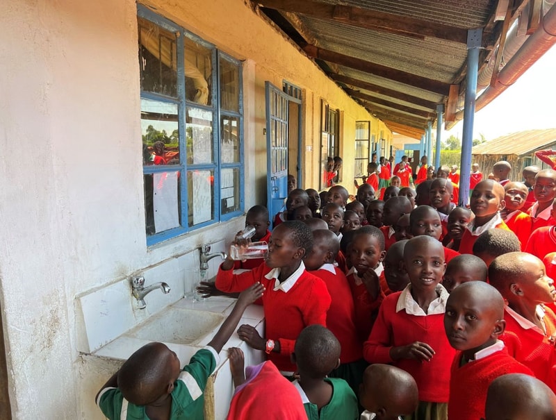 Crowded schoolyard at a drinking fountain line, many students in red uniforms gather near blue-framed windows along a sunlit corridor.