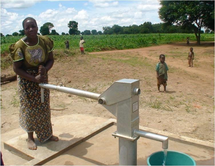 A woman stands beside a manual water pump in a rural area, with a dirt path, green fields, and a few children in the background under a partly cloudy sky.