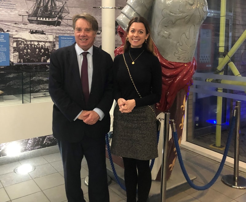 Two people stand side by side indoors, smiling for the camera, with a large statue behind them and a museum display wall in the background.