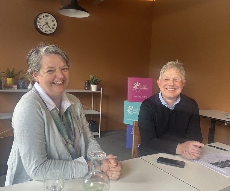 Two smiling people sit at a table in a warm room; a woman on the left wears a light jacket and scarf, and a man on the right wears a dark sweater; a bottle, glass, papers, a smartphone, and colorful boxes are on the table and behind them.