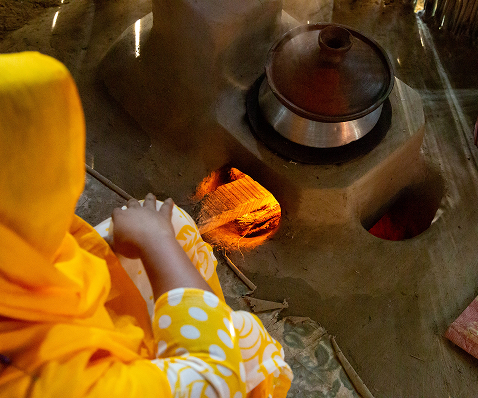 Person wearing a bright yellow headscarf and patterned clothing sits by a clay stove, tending a small fire under a metal pot.
