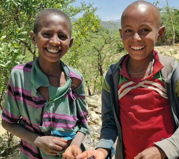 Two smiling young individuals stand outdoors in a sunny, leafy area, each holding small fruits or berries; they wear colorful striped shirts and casual jackets.