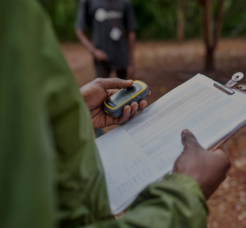 Person holds a handheld GPS device and clipboard with a printed table, outdoors, other person blurred in background, forest floor seen.