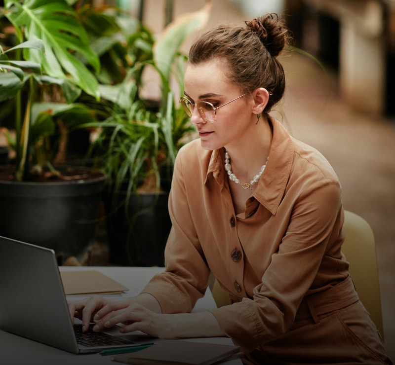 Woman with hair in a bun sits at a table typing on a laptop outdoors, wearing a tan button-up shirt, pearl necklace, and sunglasses, surrounded by green plants.