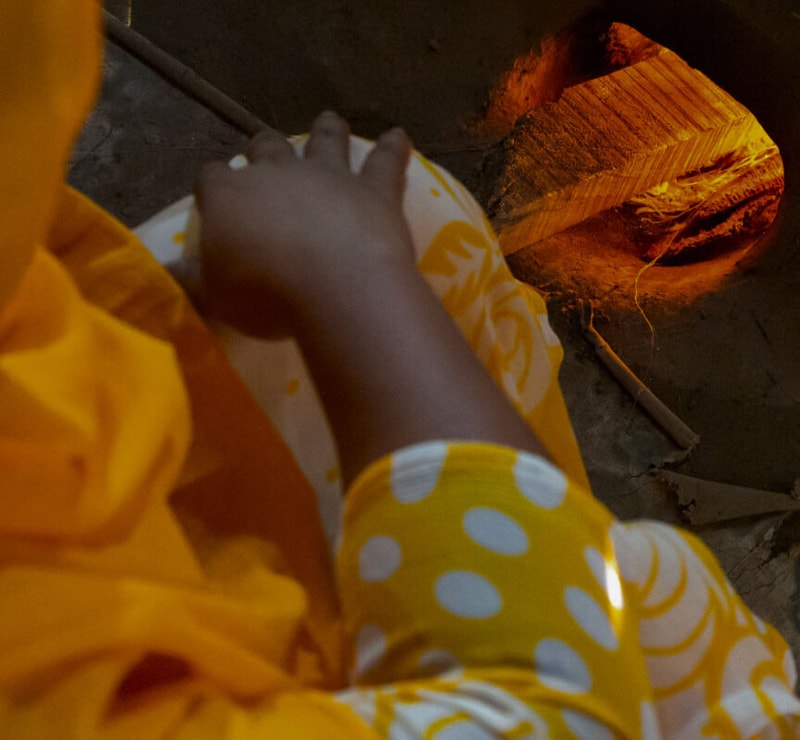 A close-up of a child wearing yellow clothing with white polka dots, resting a hand on a white fabric, near a warm orange-lit fire or kiln.