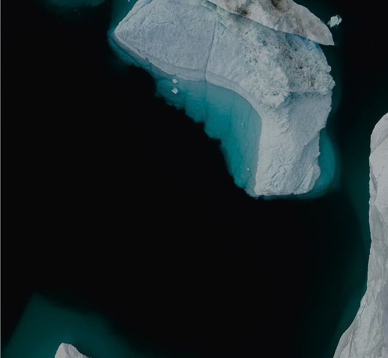 Aerial view of a dark body of water with large white icebergs along the edge, showing turquoise glow around the submerged portions of the ice.
