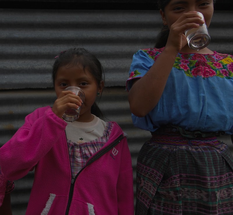 Two young girls stand against a corrugated metal wall, drinking from clear plastic cups. The left girl wears a pink fleece jacket, the right wears a blue embroidered top with a plaid skirt.