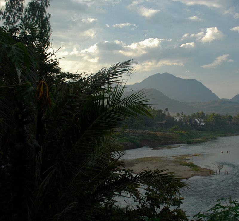 Dense tropical foliage in the foreground with a river and sandy banks curving through a valley, small houses along the shore, and hazy mountains under a cloudy sky in the distance.