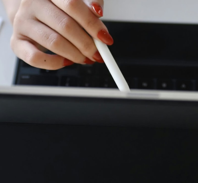A hand with red-painted nails holding a white stylus or pen and touching a device screen, with a dark keyboard-like surface in the background.