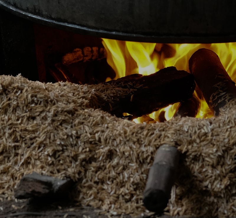 Braided sawdust and wood shavings surround a glowing wood fire inside a stove, with charred logs burning and a metal tool resting nearby.