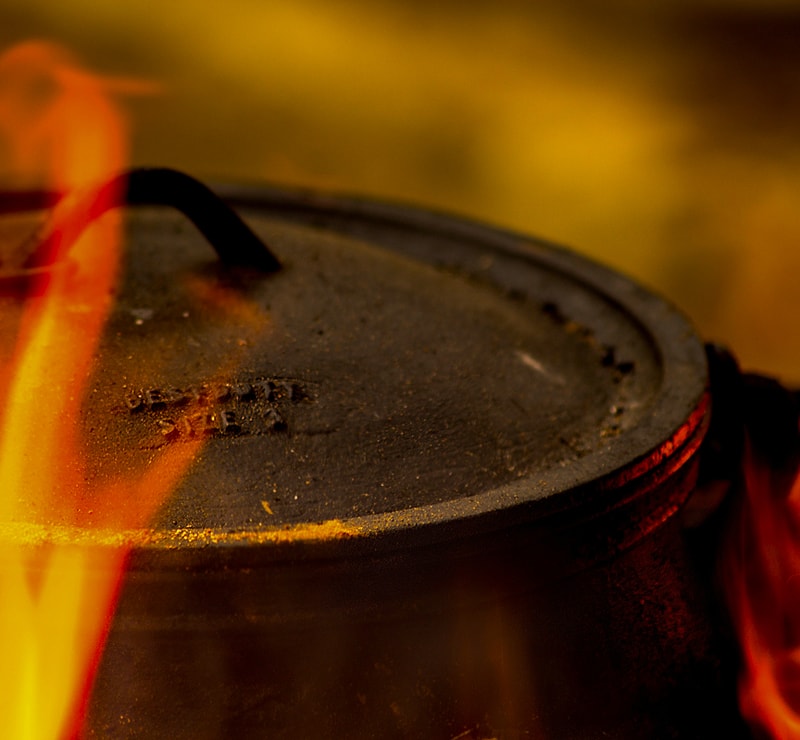A close-up of a black metal pot lid with a curved handle, surrounded by orange-yellow flames; the scene is dark and smoky with a warm glow.