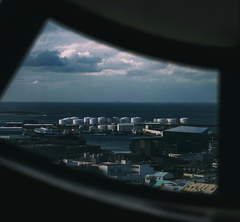 Industrial harbor scene viewed through a curved window frame, with white cylindrical storage tanks, docks, and low-rise buildings along the coastline under cloudy skies.