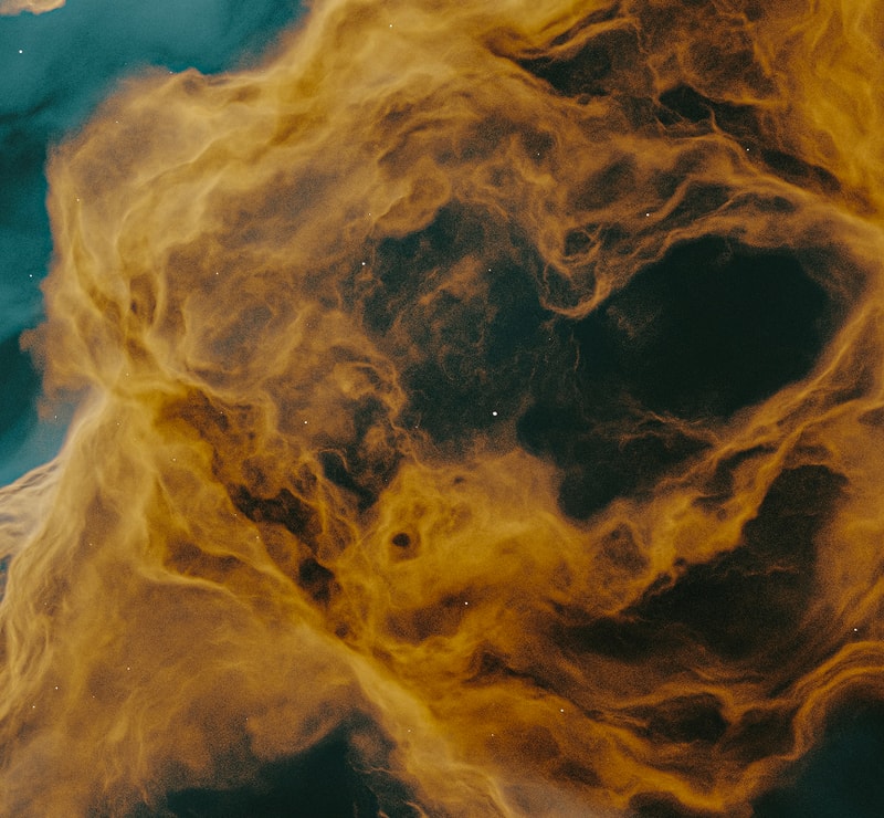 Swirling golden and orange cosmic dust forms a dense, cloudy nebula against a dark teal background, with delicate specks resembling distant stars.