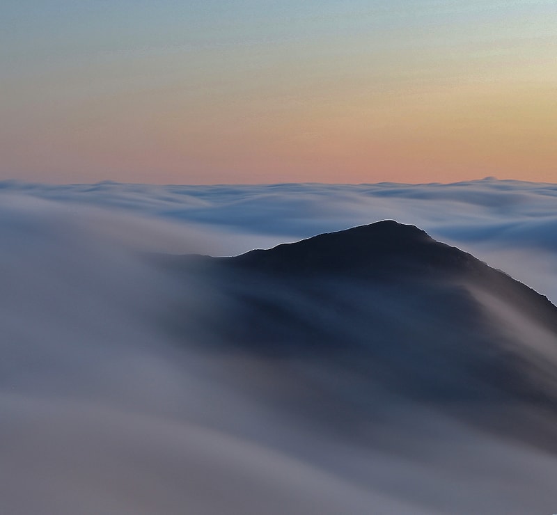 Mist-shrouded mountain peak rising above a sea of slow, wispy clouds at sunrise, with soft pastel sky gradient from pale blue to warm peach.