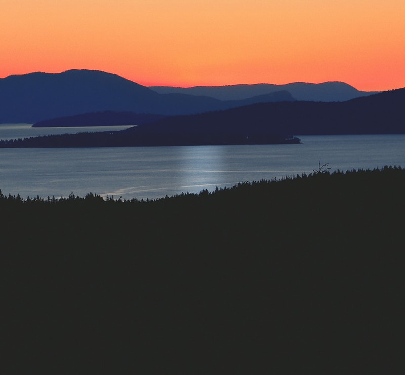 Sunset over layered silhouetted hills and a calm body of water, with warm orange sky fading to blue and a dark foreground forest edge.