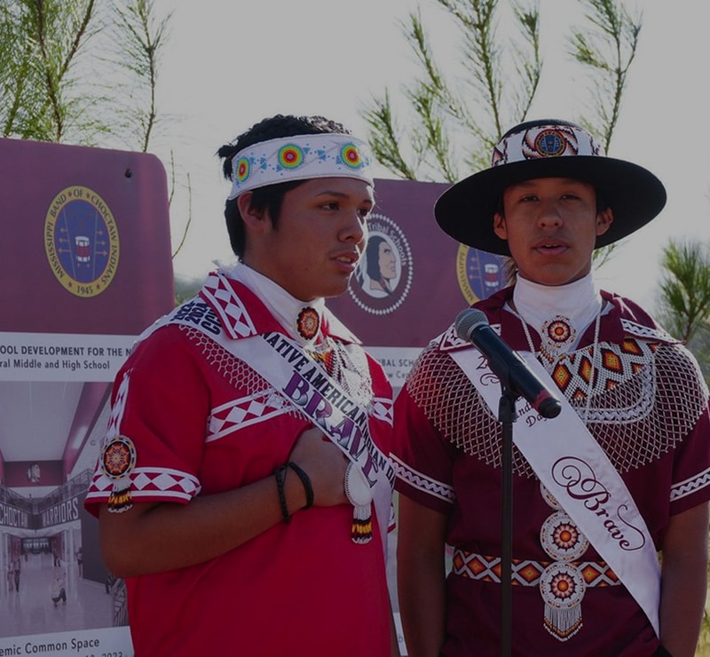 Two young people wearing colorful native-inspired outfits and sashes stand beside a microphone, outdoors, with banners in the background.