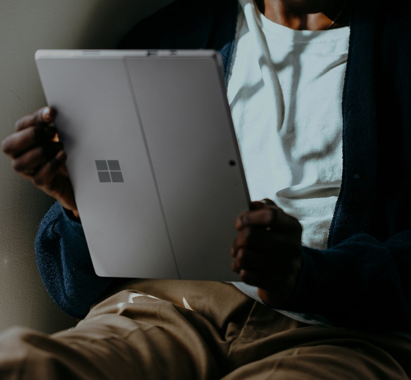 Person seated indoors holding a silver tablet with a Windows logo, wearing a white shirt and dark cardigan over beige pants.