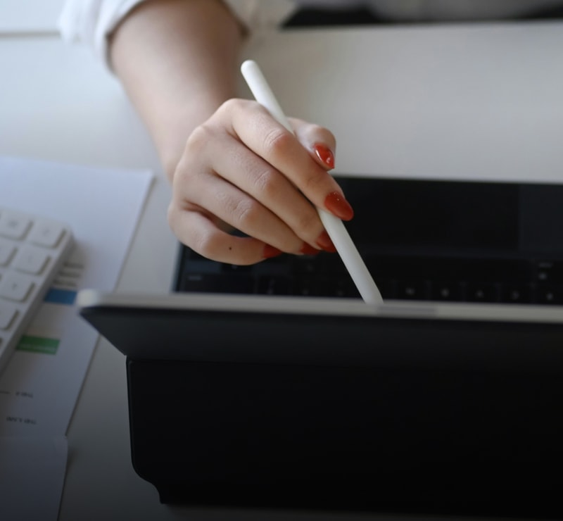 A hand with red nail polish holds a white stylus and uses a laptop touchpad, with papers and a keyboard visible on the desk in the background.