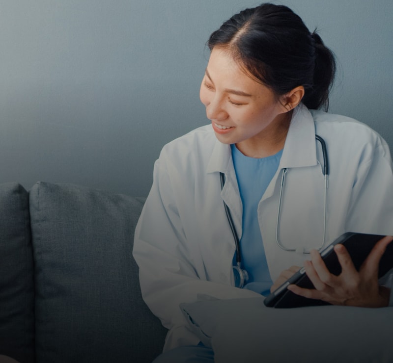A healthcare professional wearing a white coat and blue scrubs sits on a couch, smiling while looking at a tablet, with a stethoscope resting around their neck.